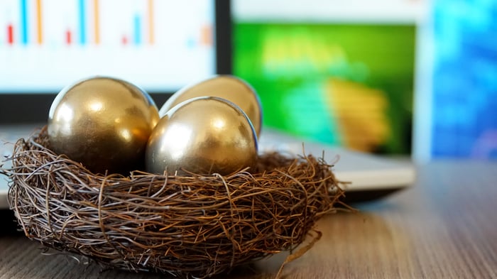 Three golden eggs in a nest in front of a laptop displaying stock charts.