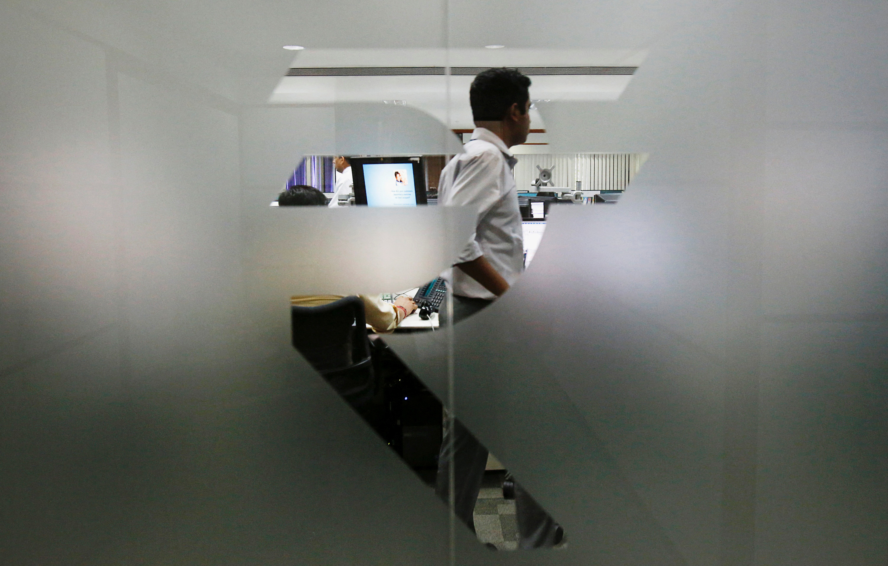 A currency trader is pictured through the symbol for the Indian Rupee on the floor of a trading firm in Mumbai