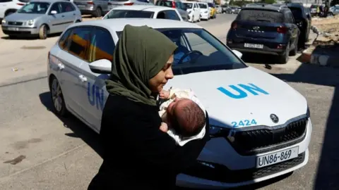 Reuters A woman carries her baby in front of a UN car at a United Nations center in Khan Younis in the southern Gaza Strip on 13 October 2023.