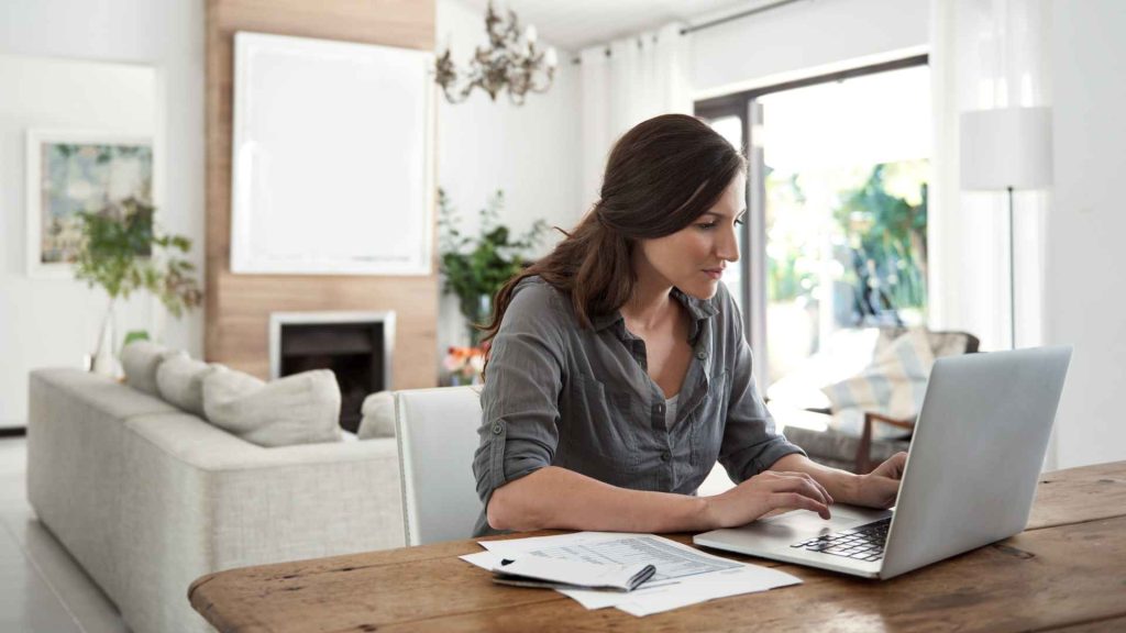 Shot of a woman using a laptop while going through paperwork at home.