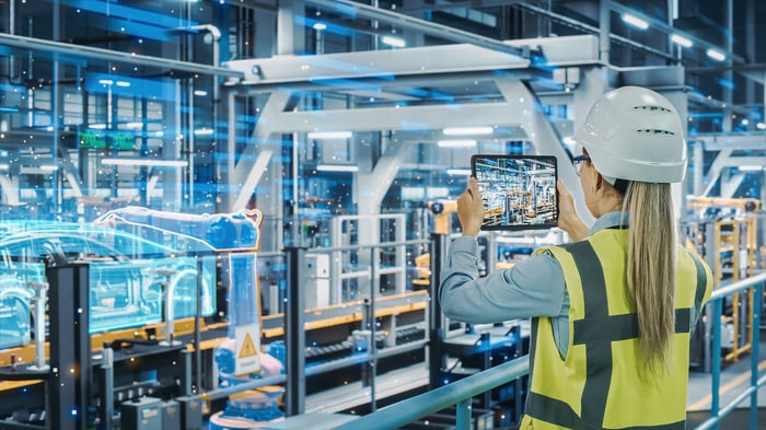 A person wearing personal protective equipment pointing a tablet at manufacturing equipment. 