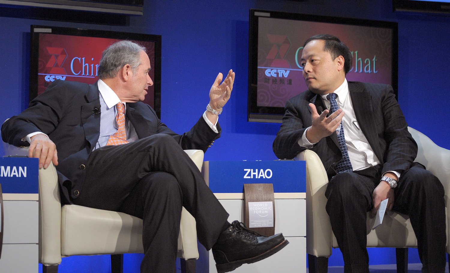 Two men sit in chairs and gesture toward each other. TV screens with CCTV logos on them are seen behind them.