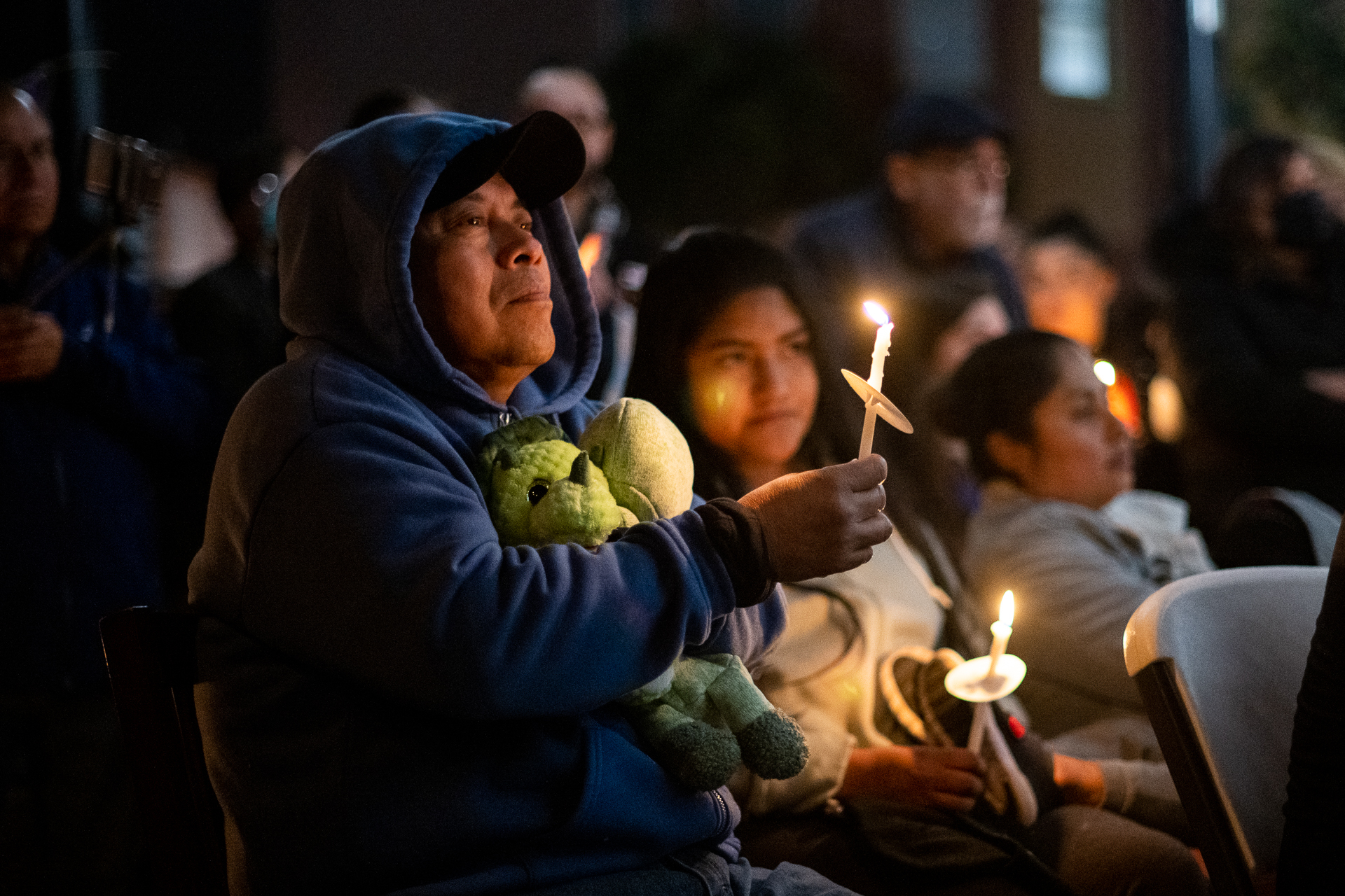 An Asian man in a blue jacket holds a candle as he looks up, with other mourners around him.