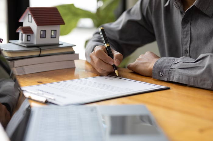 A person signs a contract with a small model home in the background.