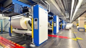 An image from inside a factory with large white and blue machines lined up next to each other, a silver ceiling with pipes and vents, red and yellow tape on the floor, and more space and pillars to the right of the machines.