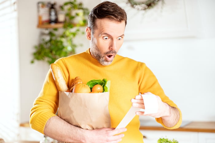 A person holds a bag of groceries and looks at a receipt with a shocked look on their face.