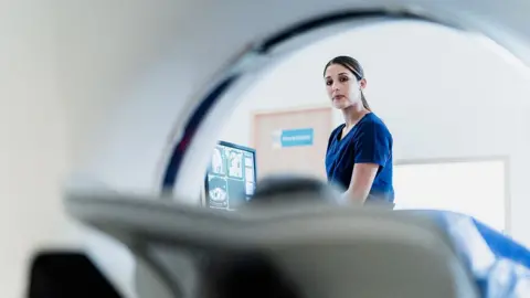 Getty Images A female healthcare professional looks over as a patient is moved into a CT scanner, the patient is out of focus and the image is taken from inside the scanner itself, with a screen showing scans and a white hospital room in the background.