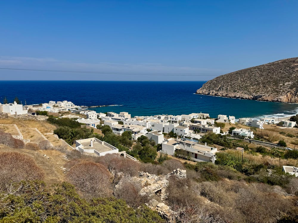 View of a coastal village with white buildings, surrounded by hills and the blue sea under a clear sky.