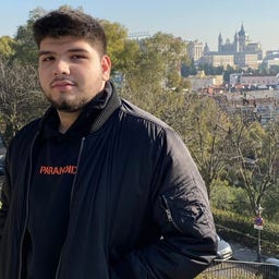 Elias Shaya looks at the camera. He stands outdoors, wearing a black jacket, with a city in the distant background.