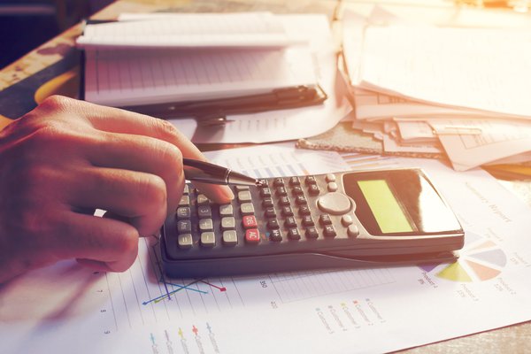 A hand using a calculator on a desk filled with paperwork.