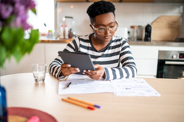 Person sitting at a kitchen table and looking at papers while holding a tablet.