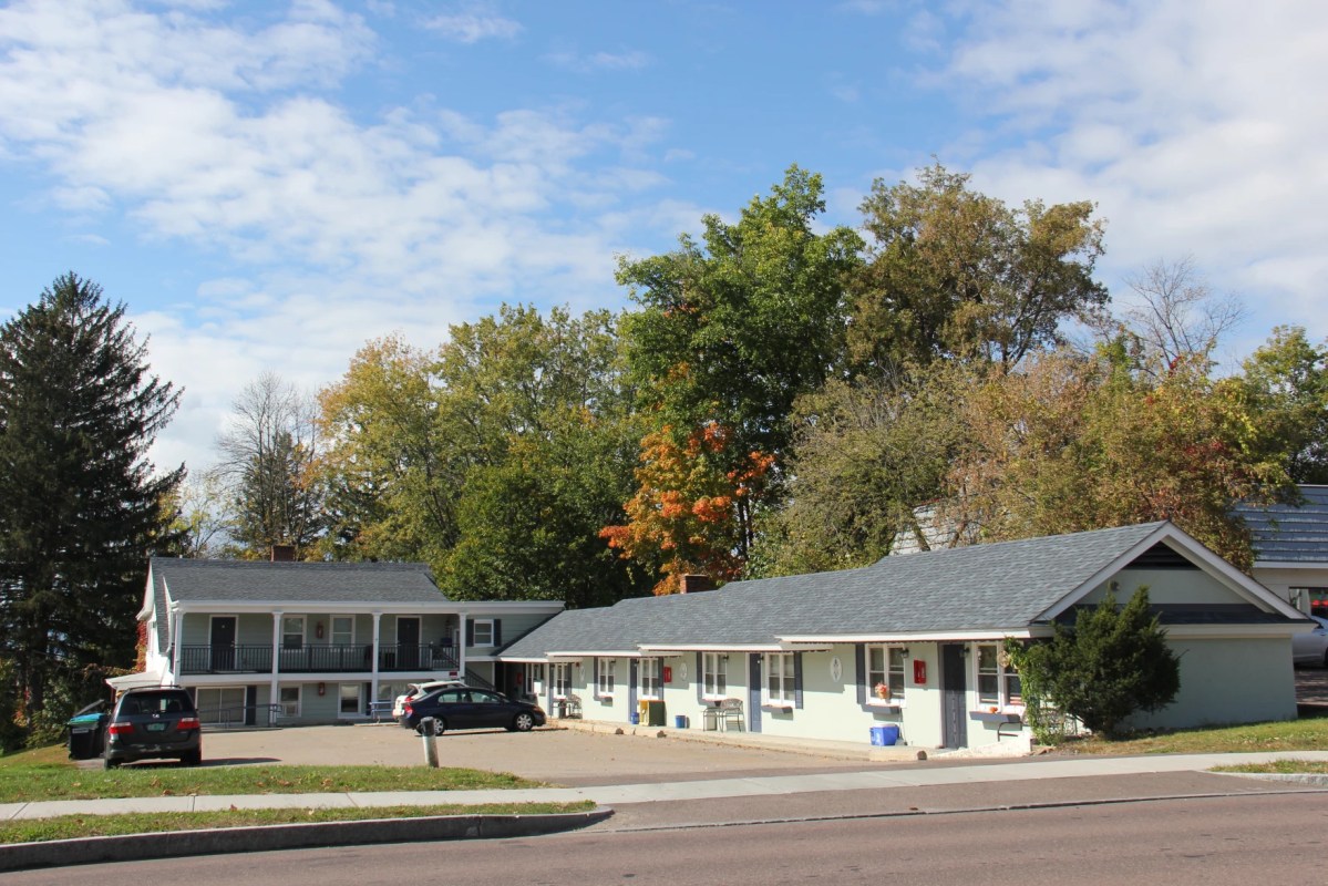 A small roadside motel with two buildings, surrounded by trees under a partly cloudy sky. Three cars are parked in the lot.