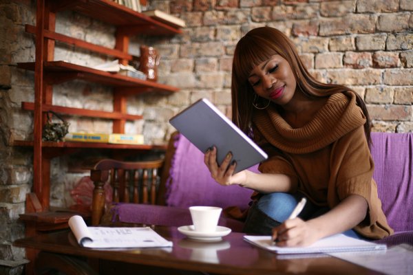 Person sitting at coffee table holding tablet and writing on notepad.