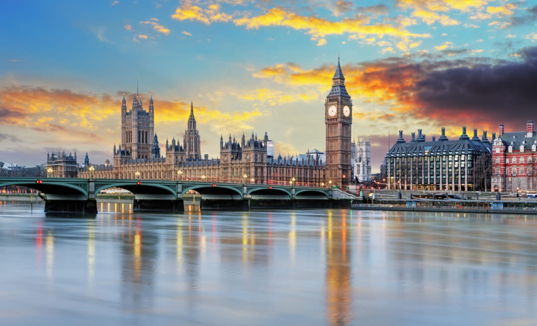 The image shows a view of the River Thames with the Houses of Parliament and Big Ben in London. The sky is vibrant with sunset colors, reflecting on the water. The historic bridge and buildings dominate the scene.