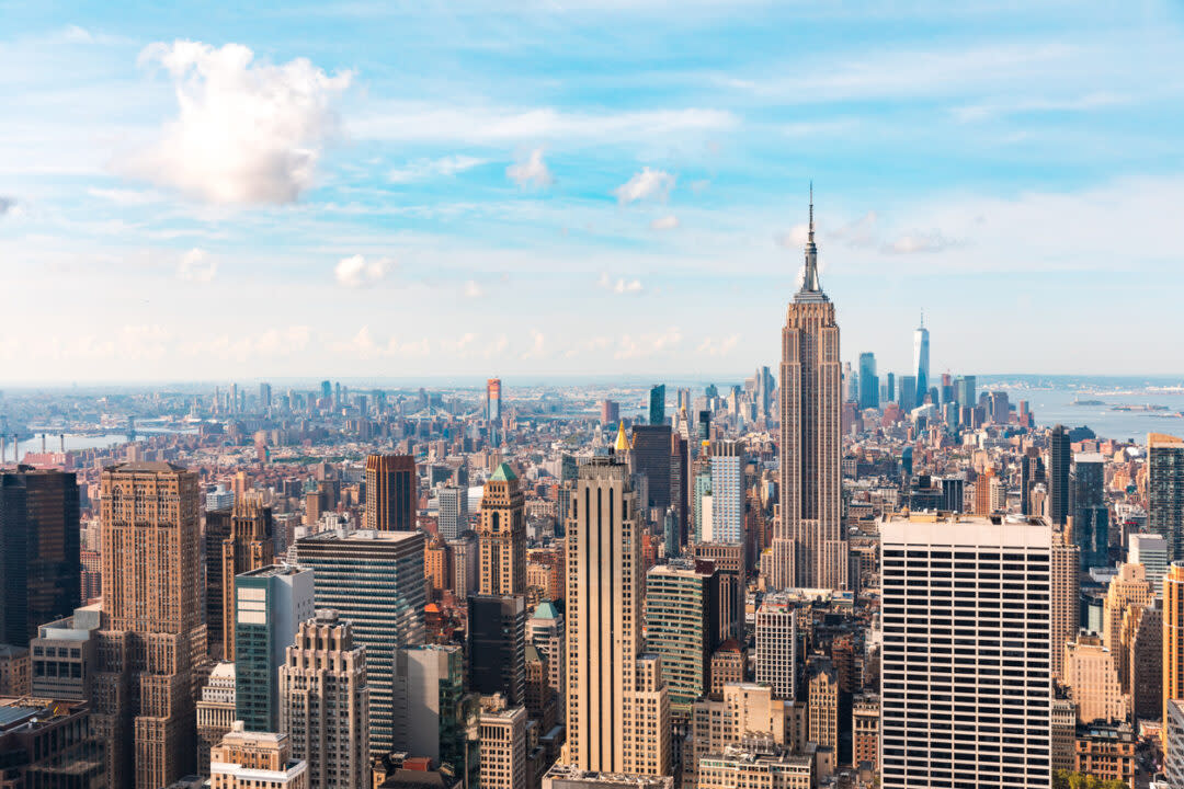 Aerial view of a city skyline featuring numerous skyscrapers under a blue sky with scattered clouds. The tallest building is situated near the center of the image, surrounded by other high-rise structures and a distant body of water.