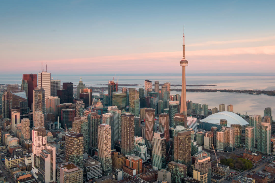 Aerial view of a city skyline with numerous skyscrapers during sunset. A prominent tower with an antenna stands near the center, and a large domed building is visible. The background features a body of water and a pastel-colored sky.