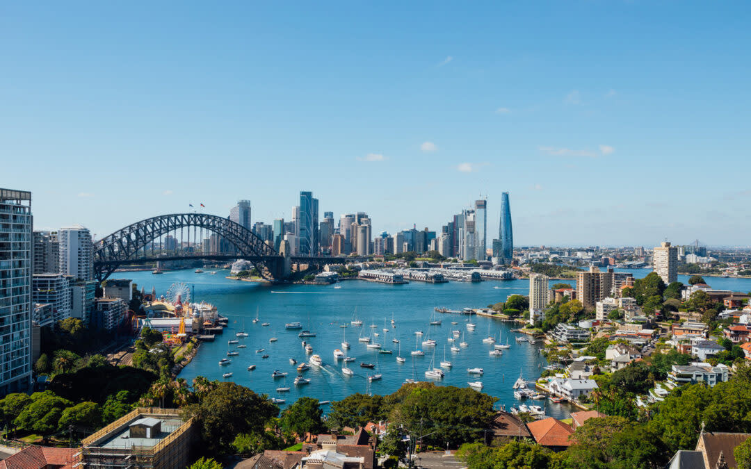 Aerial view of Sydney Harbour featuring the iconic Sydney Harbour Bridge. Numerous boats are visible in the water, with the city skyline in the background under a clear blue sky. Lush greenery and buildings are in the foreground.