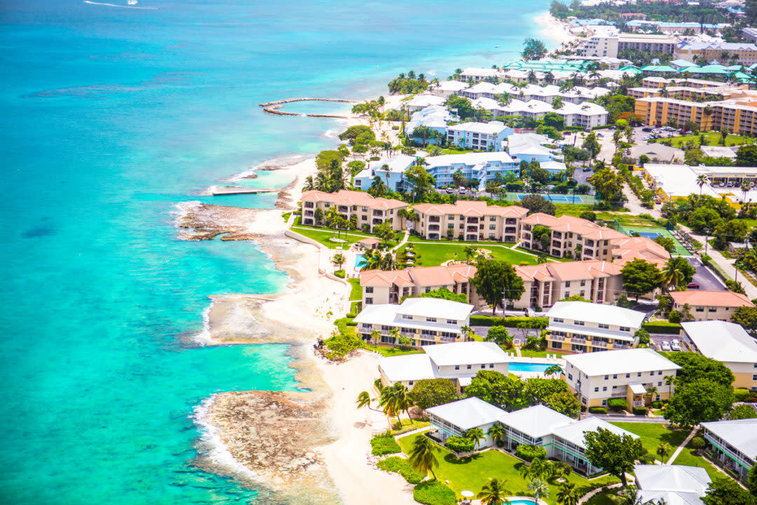 Aerial view of a coastal area with turquoise waters, sandy beaches, and a row of buildings with red roofs surrounded by greenery. Residential areas with white-roofed houses are visible further inland.