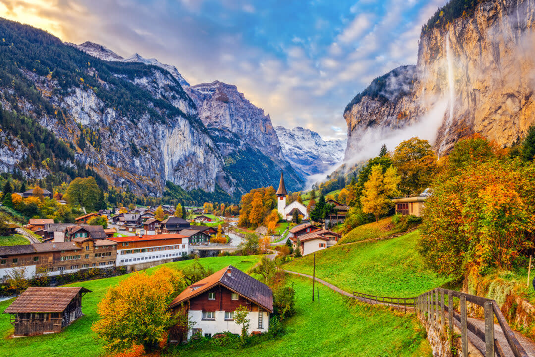 Scenic view of a Swiss village nestled in a lush valley with a river, surrounded by snow-capped mountains and autumn foliage. A church with a tall steeple sits amid charming houses, while a waterfall cascades down a cliff in the background.