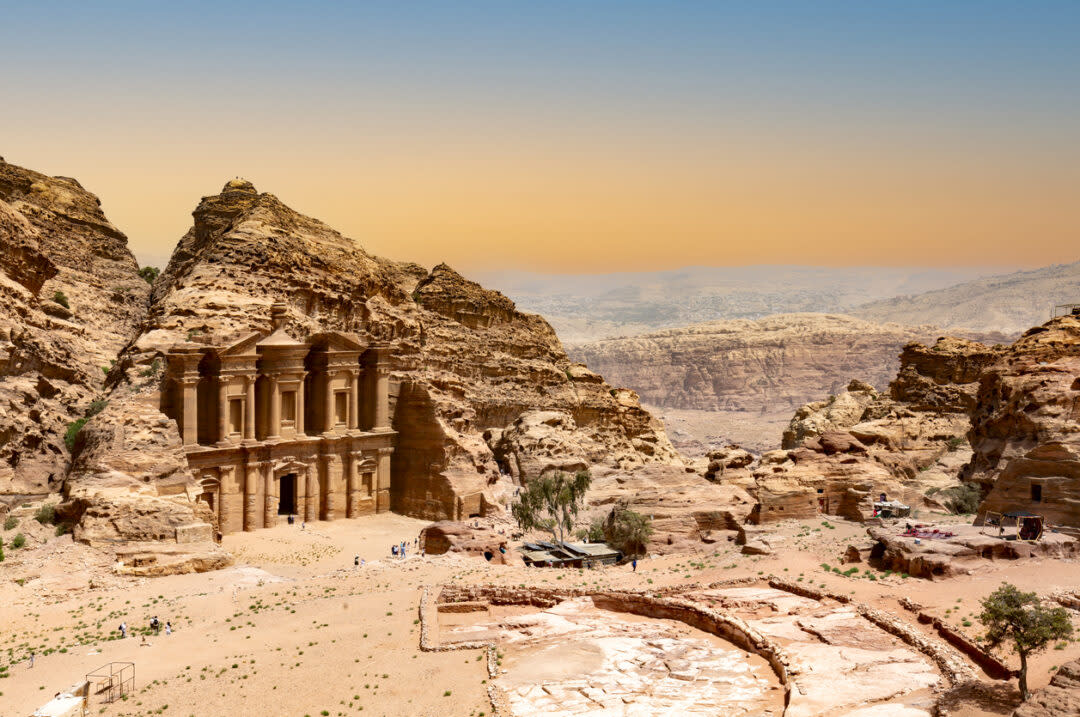 The image shows the Monastery in Petra, Jordan, a historic structure carved into sandstone cliffs. In the foreground are rocky ruins and desert vegetation, with a vast, rugged landscape in the background under a clear blue sky.