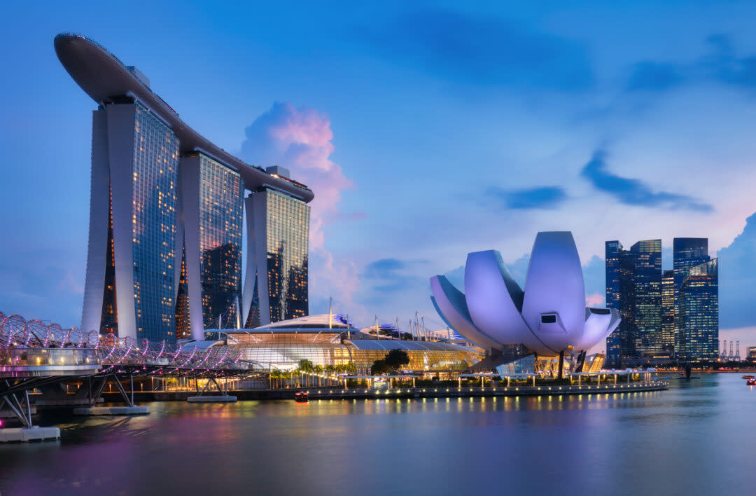 Skyline of Singapore at dusk featuring Marina Bay Sands and the ArtScience Museum. The illuminated buildings reflect on the calm water, with bright clouds and city lights enhancing the tranquil scene.