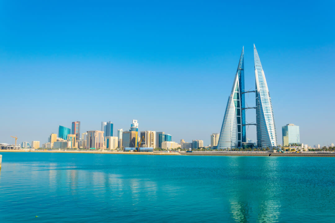 City skyline featuring the Bahrain World Trade Center with its sail-shaped towers, set against a clear blue sky. In the foreground, the calm turquoise waters of the Persian Gulf reflect the high-rise buildings.