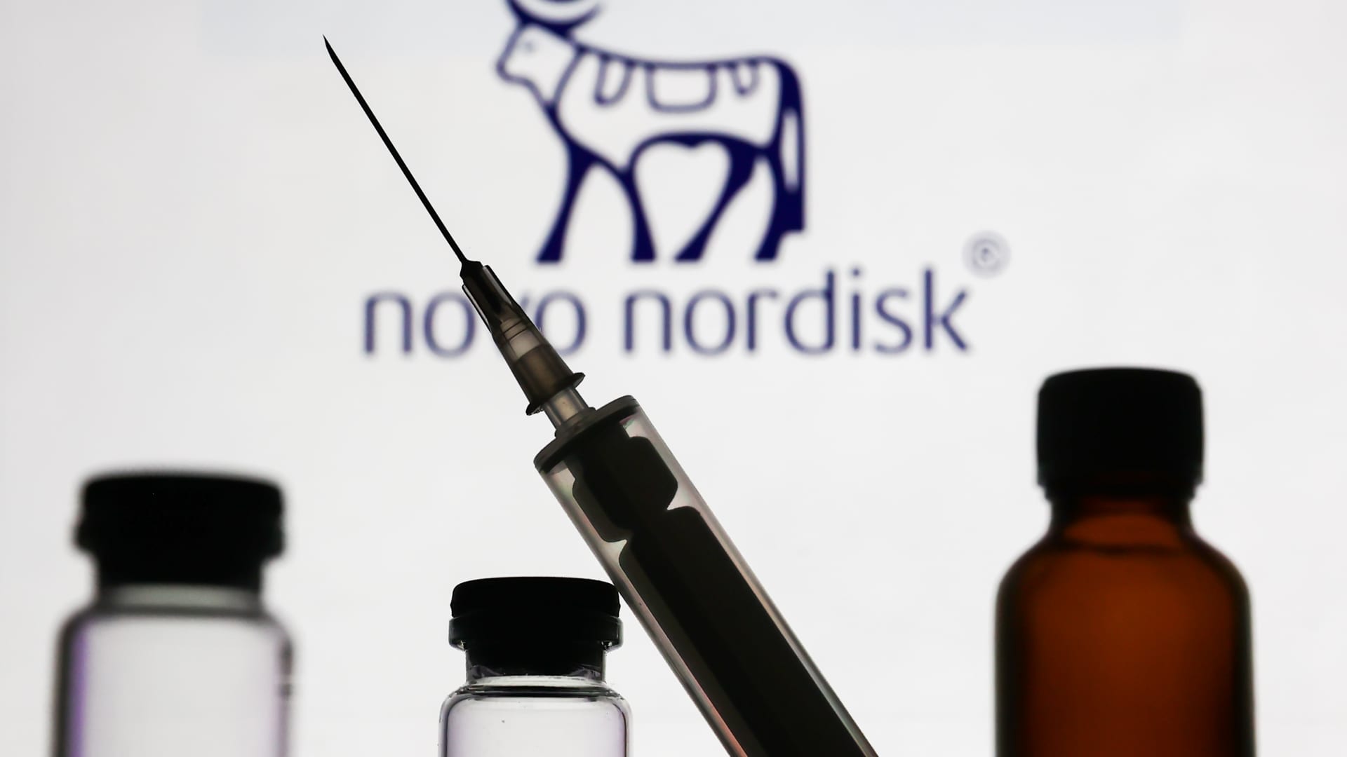 Medical bottles and syringe are seen with Novo Nordisk logo displayed on a screen in the background.