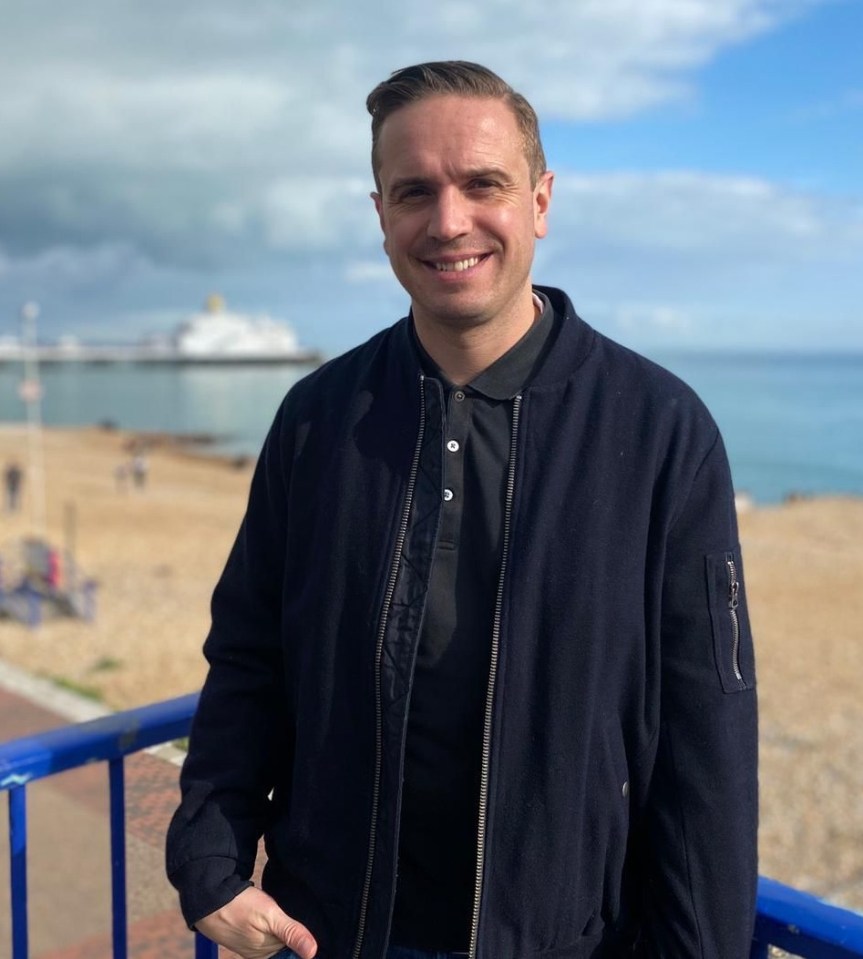 Man smiling on a beach with boats in the background.