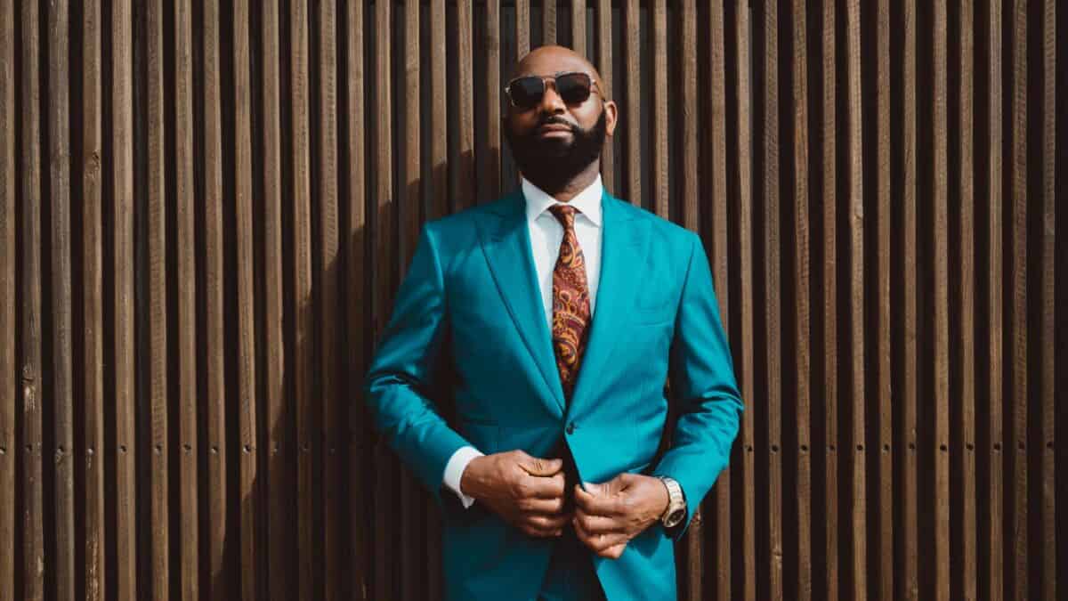 A handsome mature bald bearded black man in a sunglasses and a fashionable blue or teal costume with a tie is standing in front of a wall made of striped wooden timbers and fastening a suit button