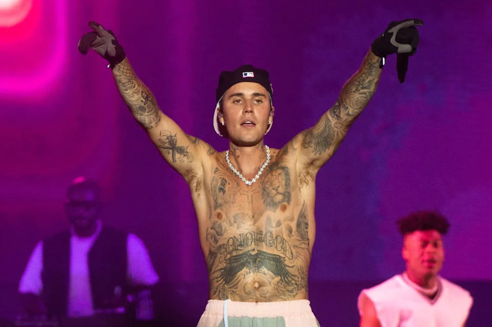 A shirtless person with tattoos performs on stage, wearing a backwards cap and pearl necklace, arms raised. Backup performers are in the background