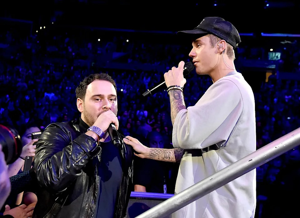 Two men perform on stage with microphones. One wears a casual sweatshirt and cap, the other a leather jacket. Audience visible in the background
