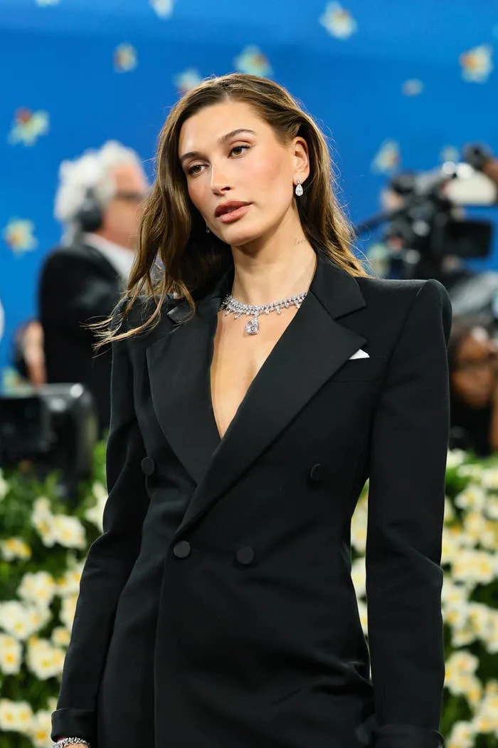 A person poses on the red carpet in a sleek black suit with a plunging neckline, accessorized with elegant diamond jewelry