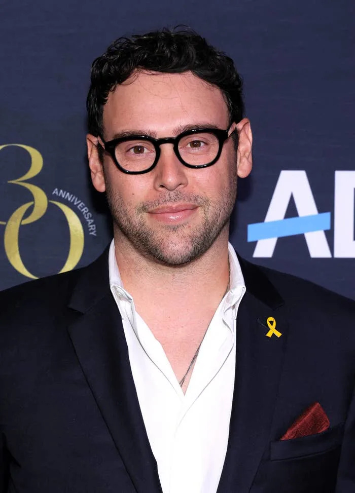 A person in a suit with a yellow ribbon pin is at an event with a dark backdrop featuring logos and the number 30