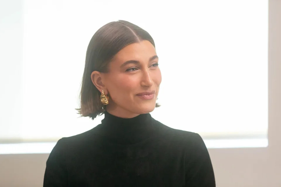 Person with short hair and gold earrings, wearing a black turtleneck, looks to the side with a slight smile