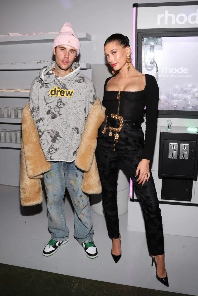 Two people posing at an event. One wears a graphic hoodie with a furry coat and jeans. The other wears an elegant black top with stylish pants