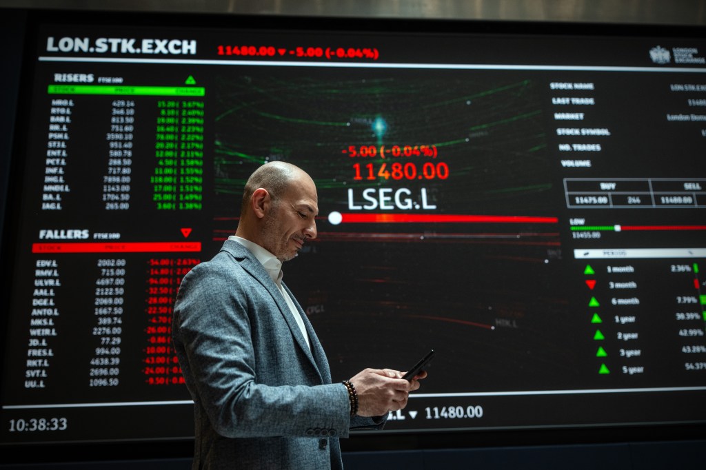 A man looks at his phone while standing in front of a stock ticker showing market data.