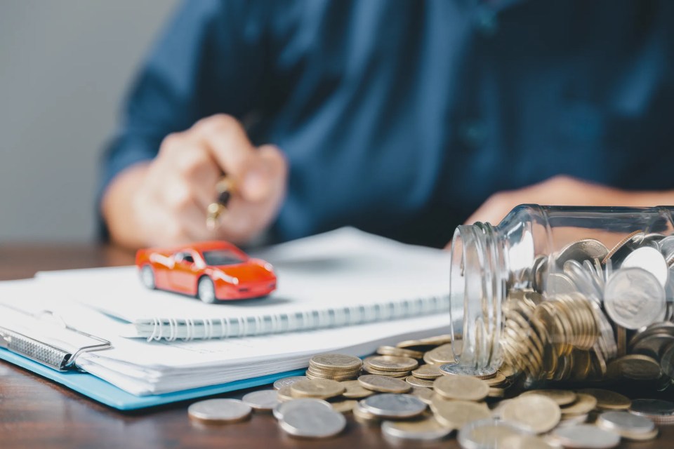 A person's hand holding a pen, with paperwork, a toy car, and a jar of coins in the foreground.