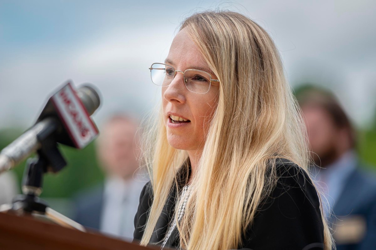 A person with long blonde hair and glasses speaks into a microphone at an outdoor event. A blurred person and green foliage are visible in the background.