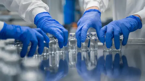 Getty Images Blue gloved hands in a lab picking up vials of clear liquid