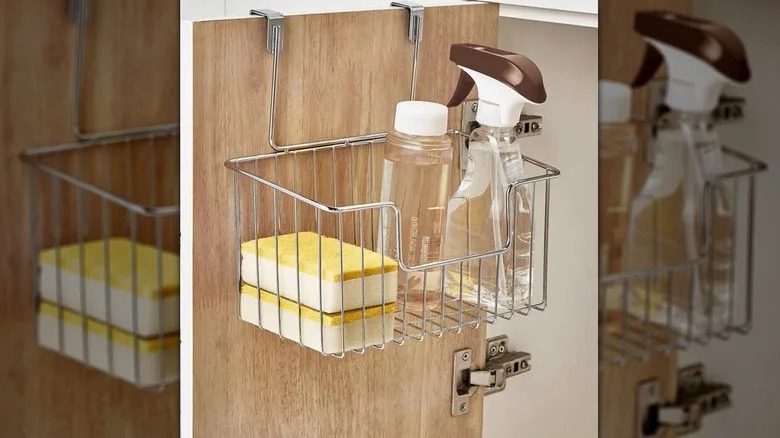 A metal hanging basket hangs over a cupboard door, containing dish sponges and bottles of cleaner