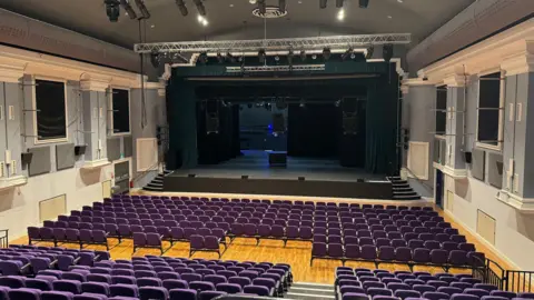 BBC Grand Hall at Dorking Halls. In the background the stage can be seen, with lighting rigs above it. In the foreground are purple coloured seats for the audience.