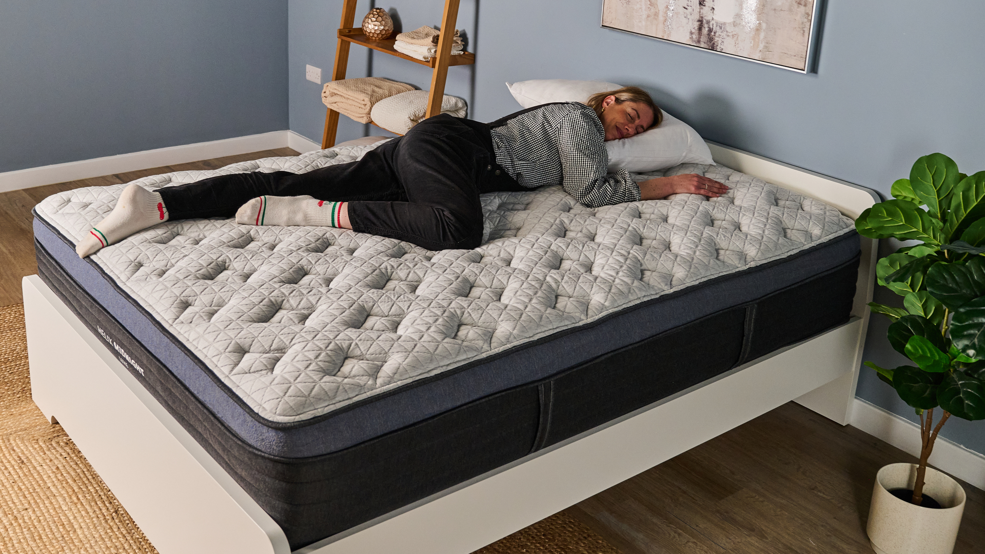 The image shows a woman wearing black dungarees lying on her side on top of a pillow top mattress