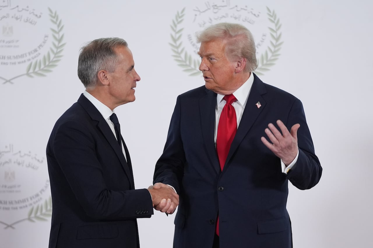 Two men in suits shake hands in front of a white screen.