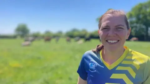 BBC A woman with brown hair and a blue and yellow patterned t-shirt in a grass field with the blurred outline of trees and animals in the background