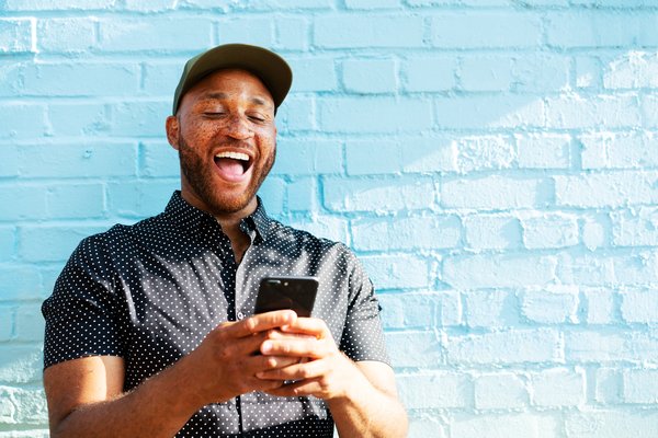 Individual wearing baseball cap smiles broadly while looking at smartphone screen outside.