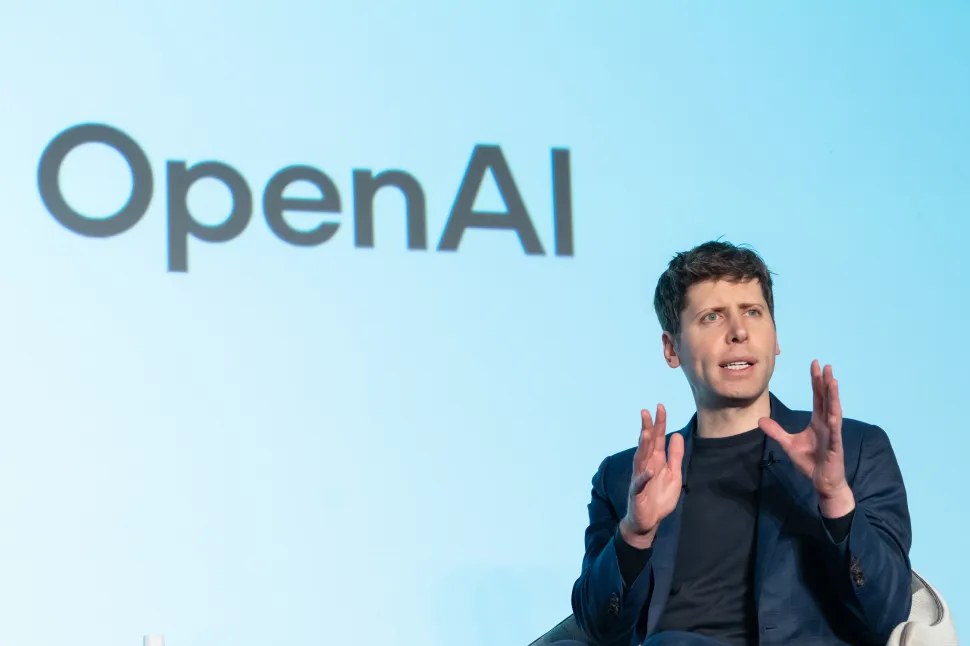 Man in black clothing sits onstage in front of blue screen reading 'OpenAI'