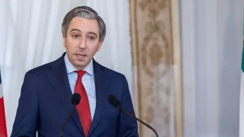 PA Simon Harris stands at a podium with two microphones. He is mid-speech. He has short, greying hair and is wearing a dark navy blue suit jacket, a blue shirt and a red tie. He is standing in front of a white wall.