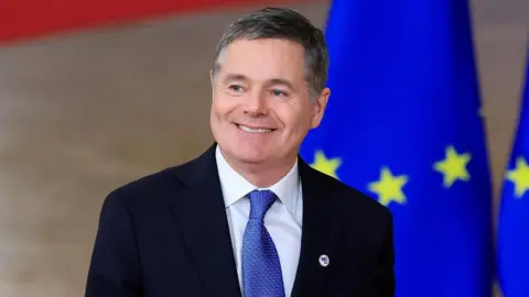 Reuters A close-up image of Paschal Donohoe attending a conference in Belgium. He is smiling at a camera. He is a middle-aged man with short, greying hair. He is wearing a dark suit jacket, a white collared shirt and a blue patterned tie.