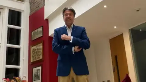 Guernsey Finance A wide shot of Rupert Pleasant. He has short dark hair and is wearing a blue suit and sandy coloured trousers.