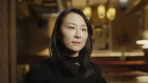 Keyu Jin is wearing a fitted black long-sleeve top with a small gold triangular pendant necklace, indoors in a warmly lit setting. The background features soft-focus hanging pendant lights with yellow shades, wooden beams across the ceiling, and muted brown walls, creating a cozy, upscale atmosphere.